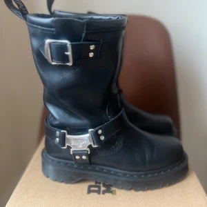 Svarta boots från Dr. Martens - Säljer ett par svarta boots från Dr. Martens med grov sula och silverfärgade spännen och detaljer. Skorna är i skinn och har en robust design med rund tå och platt sula. Perfekta för dig som gillar en tuffare stil.