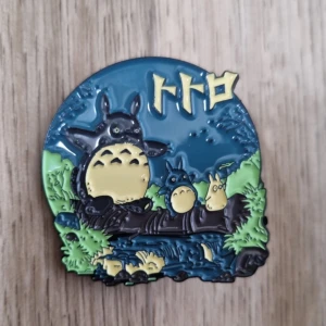 Totoro emaljpin - Säljer en färgglad emaljpin med motiv från Totoro. Pinsen har blå, grön och gul färg och japansk text upptill. Perfekt som accessoar på jacka eller väska. Storlek ca 4 cm.