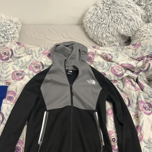 Svart och grå hoodiejacka från The North Face - Säljer en svart och grå hoodiejacka från The North Face med dragkedja och två fickor framtill. Jackan har huva och är perfekt för vardagsbruk eller utomhusaktiviteter. Klassisk logga på bröstet och ryggen.