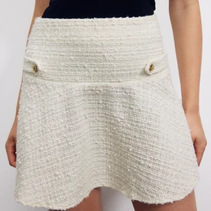 Vit tweedkjol med dekorativa knappar - Säljer en vit kortkjol i tweed med två dekorativa guldfärgade knappar framtill. Kjolen har en lätt utsvängd form och hög midja, perfekt för en chic och stilren look.