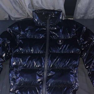 Svart pufferjacka från Trapstar - Säljer en svart glansig pufferjacka från Trapstar med hög krage och dragkedja framtill. Jackan har Trapstar-logga på bröstet och stor broderad text på ryggen. Perfekt för kalla dagar.