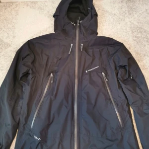 Svart vindjacka från Peak Performance - Säljer en oanvänd svart vindjacka från Peak Performance med huva och flera dragkedjeförsedda. Jackan är vartentålig och perfekt för utomhusaktiviteter.