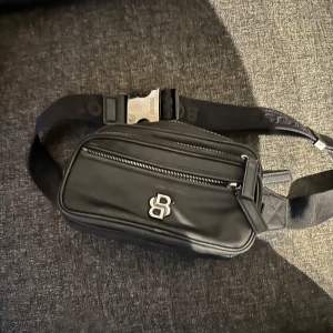 Svart axelväska från hugo boss - Säljer en svart axelväska från hugo boss med två dragkedjefack och justerbart band. Väskan har en stilren design med hugo boss framtill och passar perfekt för att bära det viktigaste.