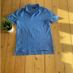 Blå pikétröja från Polo Ralph Lauren - Säljer en klassisk blå pikétröja från Polo Ralph Lauren med gul broderad logga på bröstet. Tröjan har korta ärmar, krage och knappar vid halsen. Tillverkad i 100% bomull för en skön känsla. Size 14-16 år men passar xs-s.