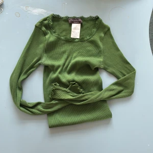 Grön ribbad topp från Rosemunde - Supersöt grön långärmad topp från Rosemunde med ribbad struktur och spetsdetaljer vid halsringningen. Perfekt för lager-på-lager och har en feminin touch med vågiga kanter vid ärmslut och hals.