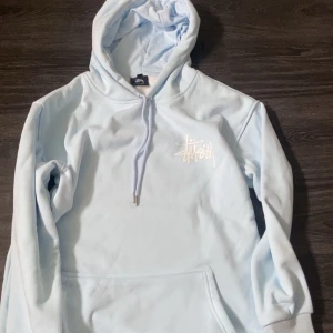 Ljusblå hoodie från Stüssy - Säljer en ljusblå hoodie från Stüssy med vit logga på bröstet och stor logga på ryggen. Tröjan har huva med dragsko och en stor magficka. Perfekt för dig som gillar streetwear och vill ha något bekvämt och snyggt.