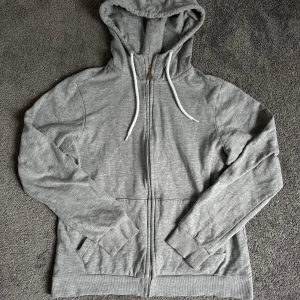 Grå zip up hoodie - Säljer en enkel och stilren grå hoodie från ETREL i storlek S. Tröjan har dragkedja framtill, huva med snören och två fickor. Perfekt för en avslappnad look och passar året runt. Koftan har ett litet hål på ryggen som har blivit igensytt, synd knappast!