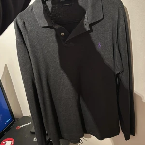 Svart pikétröja från Ralph Lauren - Säljer en stilren svart pikétröja från Ralph Lauren med lila broderad logga på bröstet. Den är i perfekt skick och inga fläckar eller liknande. Tröjan har långa ärmar och en klassisk krage. Perfekt för en avslappnad men ändå elegant look.