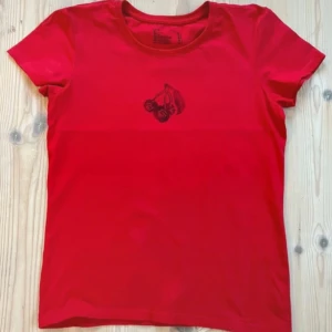 Röd t-shirt med körsbärstryck - Säljer en röd t-shirt med ett svart handprintat tryck av körsbär på bröstet. T-shirten har rund halsringning och korta ärmar. Perfekt för dig som vill ha en enkel men snygg detalj i din outfit. 