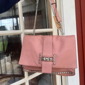 Rosa axelväska med nitar och kedja - Säljer en snygg rosa axelväska med silverfärgade nitar och kedja. Väskan har ett lock med dekorativa detaljer och flera fack inuti, inklusive dragkedja. Perfekt för dig som vill ha en cool accessoar till din outfit!