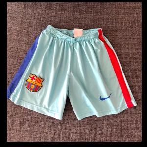 Barça träningsshorts från Nike - Ljusa turkosa träningsshorts från Nike med FC Barcelona-logga på ena benet och blå Nike Swoosh på det andra. Röd och blå rand längs sidorna. Resår i midjan för skön passform. Perfekta för fotbollsträning eller sport.