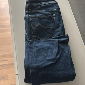 Mörkblå jeans från Jacob Cohen - Snygga mörkblå jeans från Jacob Cohen med klassisk femficksdesign och synliga knappar framtill. Jeansen har en rak passform och är tillverkade i ett mjukt denimtyg med diskreta kontrastsömmar. Perfekta för en stilren och avslappnad look.