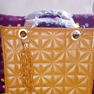 Brun quiltad handväska med kedjedetalj - Säljer en snygg brun handväska med quiltat mönster och guldfärgade metalldetaljer. Väskan har en dekorativ kedja och är perfekt för att lyfta din outfit. Handtaget är inslaget i plast för extra skydd.