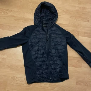 Mörkblå Peak Performance pufferjacka - Snygg mörkblå pufferjacka från Peak Performance med huva och hel dragkedja. Jackan har quiltade partier framtill, baktill och i huvan, samt släta ärmar. Perfekt för kyliga dagar och har en sportig vibe. Materialet är polyester och syntet.
