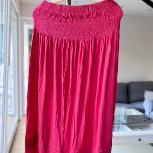 Rosa kjol - Supersöt rosa kjol av bomull med elastisk midja och skrynklig design. Den har ett flowy fall och dekorativa ränder i rosa glansig design längst ner. Perfekt för varma dagar.