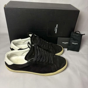  Saint Laurent skor - Snygga svarta sneakers från Saint Laurent i mocka med vit häl och gulaktig sula. Klassisk låg modell med rund tå och svarta skosnören. Guldtext på plösen och insida i vitt skinn ger en lyxig känsla. Perfekta för dig som gillar stilrena och exklusiva sneakers. ! AI BESKRIVNING! Om du har funderingar över något annat så skriv.