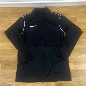 Svart Nike Dri-FIT vindjacka M - Svart vindjacka från Nike med Dri-FIT-teknologi. Jackan har hel dragkedja, vita detaljer längs axlarna och klassisk Nike-logga på bröstet. Tillverkad i lätt polyester, perfekt för träning eller chill dagar. Långärmad och sportig look.