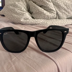 Svarta Ray-Ban Wayfarer solglasögon - Klassiska svarta Ray-Ban Wayfarer RB2140-F med bred båge och mörka glas. Snygg och tidlös design med Ray-Ban-logga i silver på skalmen. Passar perfekt om du vill ha en ikonisk look med lite edge.