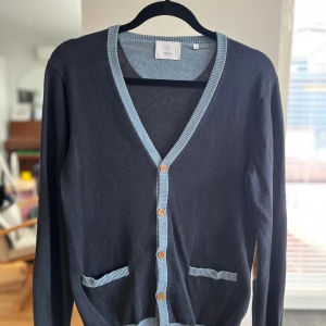 Bläck “Marcus” | Premium cardigan i 100% Pima bomull | Marinblå | S - Stilren cardigan från Bläck, modell Marcus i storlek S. Djup marinblå färg med ljusblå V-ringning och ljusbruna plastknappar. Tillverkad i 100% Pima bomull – extra mjuk, slitstark och lyxig känsla. Mått: 43 cm bred, 63 cm lång. Mycket fint skick, inga defekter. En klassisk, mångsidig kofta som passar både till vardag och jobb. Skickas snabbt och spårbart via Plick! 