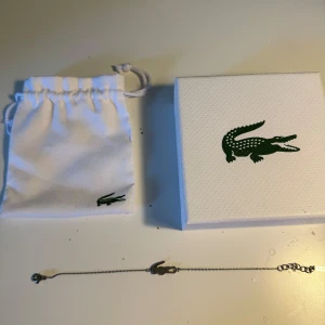 Silverfärgat armband från Lacoste - Stilrent armband i silverfärg från Lacoste med den ikoniska krokodil-loggan som detalj. Levereras med originalpåse och ask. Nypris 600kr