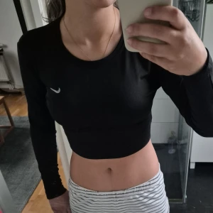 Svart croppad långärmad topp Nike - Säljer en svart croppad långärmad topp från Nike med klassisk vit swoosh-logga på bröstet. Toppen har rund halsringning och är tillverkad i ett stretchigt material som passar perfekt till träning eller sportiga outfits.