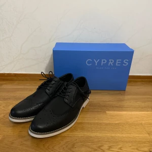 Svarta skor från Cypres med vit sula - Stilrena svarta skor från Cypres med snygga broguedetaljer och vit platt sula. Ovandelen är i skinn och textil med klassisk snörning. Perfekta för dig som vill ha en dressad men modern look.