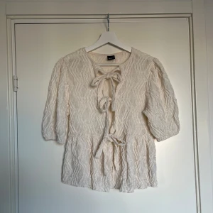 Krämvit blus från Gina Tricot - Säljer en supersöt krämvit blus från Gina Tricot med strukturmönster och knytning framtill. Blusen har volang nedtill och puffiga trekvartsärmar. 