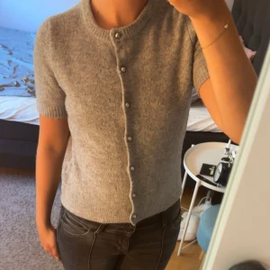 Grå kortärmad kofta med pärlknappar - Superfin grå kofta med korta ärmar och rund hals. Koftan är stickad i mjukt material och har snygga pärlknappar hela vägen framtill. 