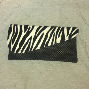 Zebramönstrad svart kuvertväska - Snygg kuvertväska i svart med zebramönstrat lock i vitt och svart. Väskan har en vävd struktur och stängs med magnetknapp. Insidan har ett mindre fack med dragkedja, perfekt för småsaker. Stilren och trendig accessoar som sticker ut.