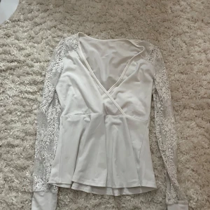 Vit omlottopp med spetsärmar - Supersöt vit omlottopp med långa spetsärmar och v-ringning. Toppen har en lätt utsvängd passform och detaljerade spetsmönster på ärmarna som ger en romantisk vibe. Perfekt att styla med jeans eller kjol för en trendig look. Står ej storlek eller vart den är ifrån 