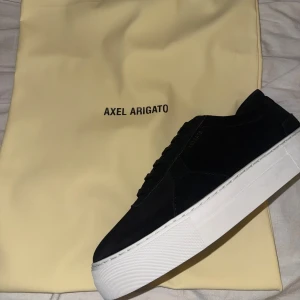 Svarta sneakers från Axel Arigato - Jag säljer nu ett par arigatos, jag fick hem de idag men kunde inte ha de. Ny pris 2700, Stilrena svarta sneakers från Axel Arigato med vit, chunky platt sula och diskret guldfärgad logga på sidan. Skorna har snörning och är tillverkade i mocka för en lyxig känsla. Perfekta för dig som gillar minimalistisk och clean stil. 