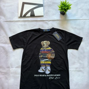 Svart Polo Bear t-shirt från Ralph Lauren - Svart t-shirt från Polo Ralph Lauren med ikoniskt Polo Bear-tryck på bröstet. Björnen har färgglad hoodie och basketboll, vilket ger en sportig vibe. T-shirten är i mjuk bomull och har klassisk passform med korta ärmar och rund hals.