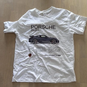 Vit Porsche 911 GT3 t-shirt - Vit t-shirt med stor Porsche 911 GT3-print på bröstet och tydlig Porsche-logga. Klassisk passform med rund hals och korta ärmar. Perfekt för dig som älskar bilar och vill sticka ut med en snygg och sportig look.