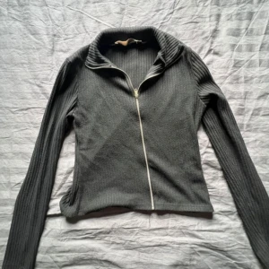 Svart ribbad kofta med dragkedja - Snygg svart ribbad kofta med lång ärm och hel dragkedja framtill. Tröjan har en hög krage och är figurnära i modellen, perfekt att slänga på sig när det är lite kyligt. Enkel och stilren design som funkar till det mesta. Från lager 157 storlek M !💕🙌