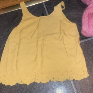 Gul linnetopp med broderad kant - Söt gul linnetopp med breda axelband och broderad, vågig kant nertill. Perfekt för varma dagar och ger en fräsch look med sin ljusa färg och fina detaljer. Toppen är ärmlös och har en luftig passform.