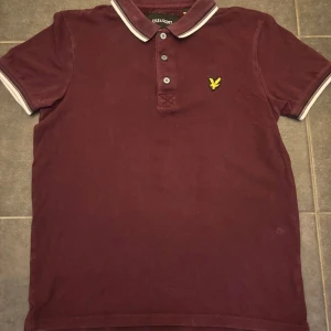 Vinröd pikétröja från Lyle & Scott - Snygg vinröd pikétröja från Lyle & Scott med klassisk krage och tre knappar framtill. Tröjan har vita detaljer på krage och ärmslut samt den ikoniska gula loggan på bröstet. Perfekt för en stilren och avslappnad look.