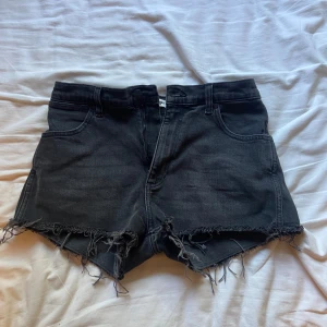 Svarta jeansshorts med fransig kant - Snygga svarta jeansshorts från Gina Tricot med hög midja och rå, fransig kant nertill. Klassisk femficksmodell med dragkedja och knapp. Perfekta för sommaren och ger en cool vibe till din outfit.