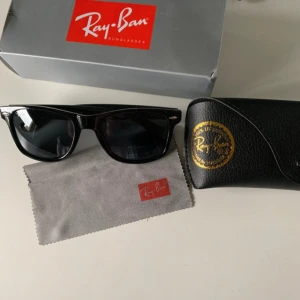 Svarta Ray-Ban Wayfarer solglasögon - Klassiska svarta Ray-Ban Wayfarer solglasögon med snygg, tidlös design. Bågarna är i blank svart plast och glasen är mörka. Kommer med originalfodral, putsduk och box. Perfekta för dig som vill ha en ikonisk look.