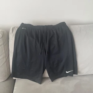 Svarta Nike Dri-Fit träningsshorts - Svarta träningsshorts från Nike med Dri-Fit-teknologi. De har elastisk midja med snörning och en klassisk loose passform. Perfekta för gymmet eller löprundan, med diskret Nike-logga i vitt på ena benet.