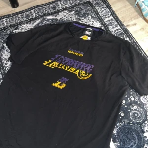 Hugo BOSS NBA - Svart t-shirt från BOSS x NBA med Los Angeles Lakers tryck i lila och gult på bröstet. Klassisk rund hals, kort ärm och coolt Lakers-märke både fram och i nacken. Perfekt för dig som älskar basket och streetstyle. Går knappt att få tag på längre…