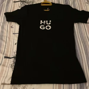 Svart t-shirt från HUGO med tryck - Svart t-shirt från HUGO med vit grafisk logga på bröstet. Klassisk passform med korta ärmar och rund halsringning. Tillverkad i mjuk bomull som känns skön mot huden. Perfekt för dig som gillar stilrena och trendiga plagg med tydlig branding. Perfekt tröja från Hugo boss som går att använda dagligen. Den är helt ny och priset kan diskuteras hör av vid frågor och funderingar!