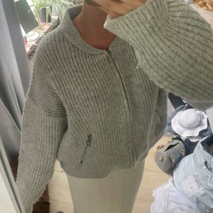 Ljusgrå stickad cardigan tröja - Mysig ljusgrå stickad cardigan tröja med grov ribbad struktur och dragkedja framtill. Jackan har två fickor med dragkedjor och en rund halsringning. Perfekt för lager-på-lager och chill vibes.
