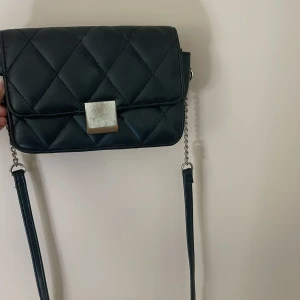 Svart quiltad axelväska med kedja från glitter - Snygg svart axelväska från glitter med quiltat mönster och silverfärgad kedjerem. Väskan har en fyrkantig metallknapp framtill och ett innerfack med dragkedja. Perfekt storlek för det viktigaste. Säljs i befintligt skick som syns på bilden! 