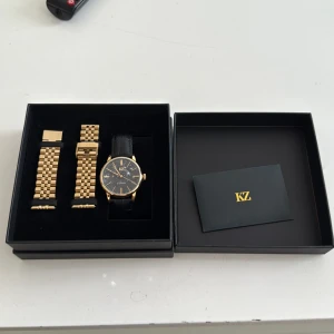 Svart och guld klocka från KZ - Stilren klocka från KZ med svart urtavla och guldfärgade detaljer. Kommer med både svart läderarmband och ett extra guldfärgat metallarmband. Levereras i original ask med KZ-logga. Perfekt accessoar för dig som gillar klassisk och lyxig stil.