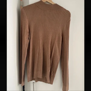  polotröja - Säljer en beige ribbad polotröja med lång ärm. Tröjan har en högre hals och är tillverkad i ett mjukt, stretchigt material som sitter snyggt på kroppen. Perfekt för lager-på-lager