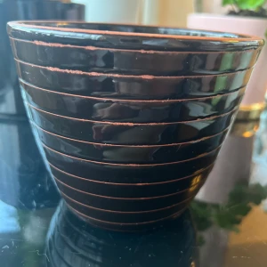 Kruka  - Bilderna visar en svart kruka med horisontella ränder och terrakottafärgad kant. Produkten är märkt 'Krukka Domino Slat-Rand SV. 15 cm' från R&A Collection