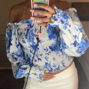 Blommig blus från Gina Tricot - Supersöt blommig blus från Gina Tricot i vitt och blått. Toppen har smockad kropp, långa puffiga ärmar med resår och offshoulder-modell. Perfekt för dig som gillar romantiska vibes och vill sticka ut med ett fräscht mönster.