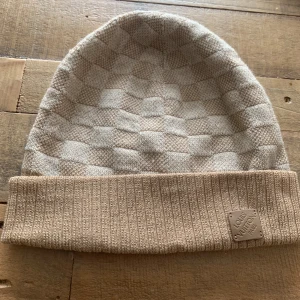 Beige rutig mössa från Louis Vuitton - Exklusiv beige mössa från Louis Vuitton med rutigt mönster och ribbad uppvikt kant. Tillverkad i 100% ull med en patch i kalvskinn framtill. Snygg och stilren accessoar som lyfter din look.    Har kvitto och låda som följer med där du kan se köpet en beige mössa 