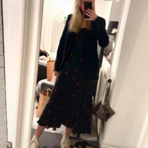 Adoore Verona klänning - Supersnygg svart adoore klänning med tunna axelband, öppen rygg med snörning och små röda blommor. Klänningen har en figurnära passform och faller snyggt längs kroppen. Perfekt för festliga tillfällen 💗