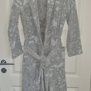 Blommig badrock  från Pelle Vävare stl xs - Snygg badrock ii frotté från Pelle Vävare Sweden med grått och beige blommönster. Badrocken har knytskärp i midjan och bred krage. Perfekt för att chilla hemma eller efter duschen, med en riktigt mjuk och skön känsla.